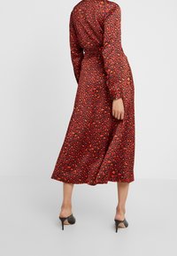 Femme portant une robe midi à manches longues imprimé léopard rouge et noir avec une ceinture à la taille, associée à des mules noires à talons hauts.