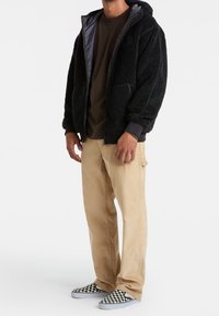 Schwarze, fleecegefütterte Kapuzenjacke mit grauen Akzenten, kombiniert mit beigen Hosen und schwarz-weißen karierter Slipper.