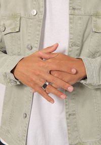 Grön denimjacka med silverdetaljer, bärs över en vit skjorta. Armarna är korsade och visar en tvåfärgad ring med en rektangulär svart sten.