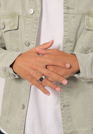 Grøn denimjakke med sølvhardware, båret over en hvid skjorte. Hænderne er krydset, og viser en tofarvet ring med en rektangulær sort sten.