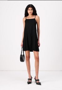 Vestido preto sem mangas, de corte reto, feito de tecido leve. Combinado com sapatos de salto alto pretos e uma pequena bolsa.