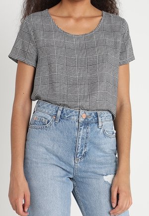 Femme portant un haut à manches courtes à carreaux noir et blanc rentré dans un jean déchiré bleu clair taille haute, debout devant un fond uni.
