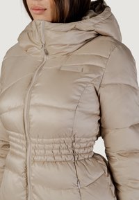 Giacca a puffo beige chiaro con un design aderente, cappuccio, sezioni trapuntate verticali e vita elasticizzata. Presenta un logo discreto sul petto.