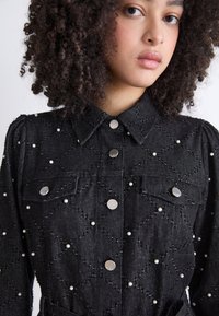 Camicia di denim nera con motivo trapuntato, decorata con perle bianche e bottoni a scatto argentati. Collo classico, tasche frontali e maniche a sbuffo.