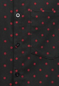 Tecido preto com bolinhas vermelhas apresenta um bolso frontal no peito e uma fileira vertical de botões pretos e brancos.