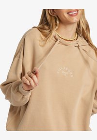 Sudadera de color beige claro, de tela suave, corte holgado, con capucha ajustable. Presenta un logo circular blanco que dice "BILLABONG SINCE 73".