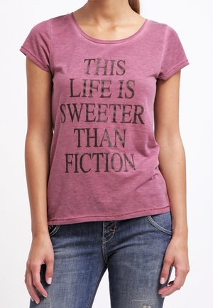 Mujer llevando una camiseta púrpura desgastada con el texto "THIS LIFE IS SWEETER THAN FICTION" y vaqueros azules, de pie con los brazos relajados.