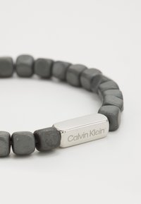 Grau-perlenarmband mit quadratischen Perlen und einem rechteckigen silbernen Metallakzent, der mit "Calvin Klein" graviert ist. Glatte Textur, matte Oberfläche.