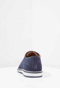 Chaussure en daim bleu marine avec un dos arrondi, un intérieur marron contrastant et une semelle en caoutchouc blanche rayée. Texture lisse, design minimaliste.