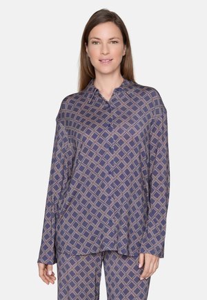 Femme aux cheveux bruns lisses portant une chemise ample violette à motifs avec boutons et un pantalon assorti sur un fond clair uni.