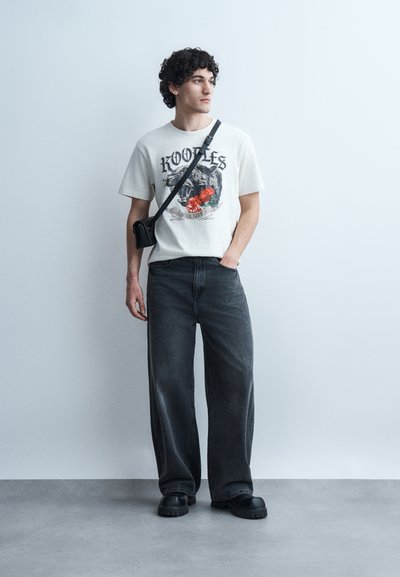 Νεαρός άνδρας που στέκεται, φορεί ένα λευκό γραφικό μπλουζάκι, φαρδιά μαύρα τζιν, μαύρα chunky παπούτσια και μια μαύρη τσάντα ώμου, κοιτάζοντας πλάγια.