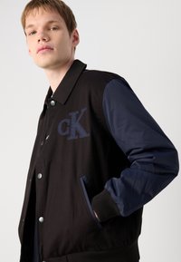 Black fabric jacket with blue sleeves, featuring snap buttons, side pockets, and embroidered "CK" logo on the chest.