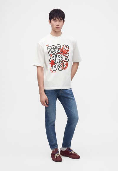 Jeune homme portant un t-shirt graphique blanc avec un texte noir et rouge, un jean bleu et des baskets rouges, debout devant un fond uni.