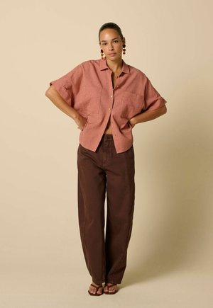 Femme debout, mains sur les hanches, portant une chemise rose ample à manches courtes texturée, un pantalon marron à jambes larges et des boucles d’oreilles en forme de cœur.