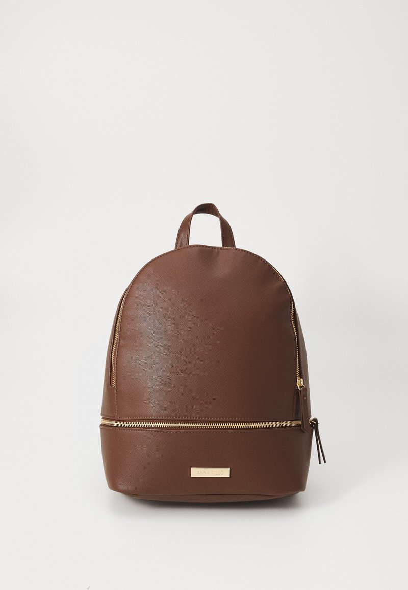 Sac à dos en cuir synthétique marron avec un dessus arrondi, quincaillerie dorée et une poche frontale zippée. Il dispose d'une poignée supérieure pour le transport.