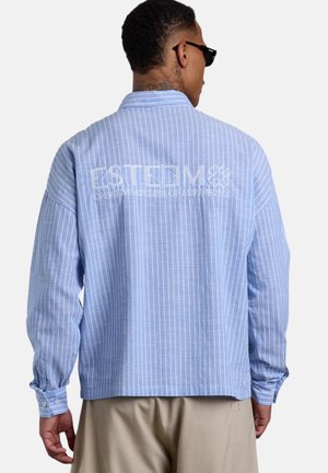 Mann trägt ein hellblaues, weiß gestreiftes Langarmhemd mit Text und Logo auf dem Rücken, beige Hose und schwarze Sonnenbrille.