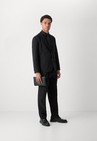 Black suit with a fitted jacket and straight-leg trousers. Paired with a black shirt, tie, and leather shoes. Holding a black clutch.