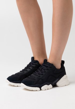 Legs wearing black lace-up sneakers with chunky white soles, standing on a plain white surface.