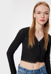 Musta lyhythihaisten yläosien cropped-malli, jossa on ribbitekstuuri. Yläosassa on pitsireunus kaula-aukon ja helmarenkaan ympärillä, sekä etupuolella napit kiinnitystä varten.
