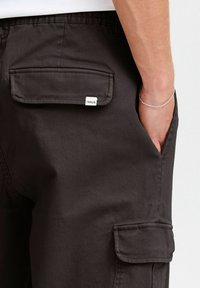 Schwarze Cargo-Hosen aus glattem Material, mit flachen Taschen auf der Rückseite und an den Seiten, versehen mit einem kleinen Etikett mit der Aufschrift "SOLID".