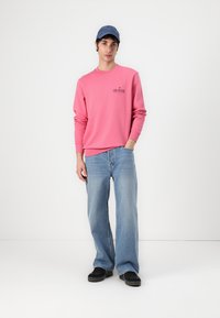 Rosa genser med ribbet mansjetter, blå bukser med vide ben, og sorte joggesko. En blå caps er på hodet. En enkel logo detalj på genseren.