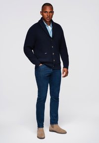 Cardigan lavorato a maglia blu navy con colletto a scialle, chiusura con bottoni e texture a coste, abbinato a jeans blu scuro e stivaletti in suede beige chiaro.