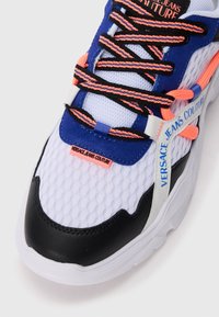Białe siatkowe sneakersy z niebieskimi i czarnymi akcentami, pomarańczowymi sznurowadłami i czarnym noskiem; metka z logo na boku z napisem "Versace Jeans Couture."