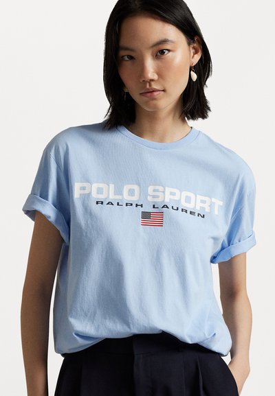 Világoskék pamut póló, felgöngyölt ujjakra jellemző, nagy fehér "POLO SPORT" szöveggel, "RALPH LAUREN" felirattal és egy kis amerikai zászló grafikával.