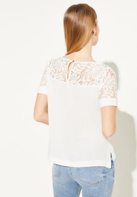 Haut blanc à manches courtes avec un empiècement en dentelle, fermeture en forme de trou de serrure au dos et ourlet droit. Tissu doux avec une texture lisse, coupe ajustée.