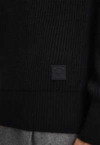 Maglione nero a coste con finitura testurizzata e una toppa quadrata nera vicino all'orlo. Presenta una vita aderente e maniche lunghe.