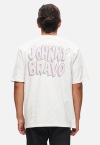 Bílý bavlněný tričko s velkým růžovým grafickým nápisem "JOHNNY BRAVO" na zádech. Krátké rukávy, volný střih a design s kulatým výstřihem.