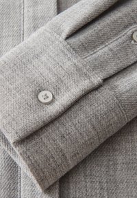 Cuff di camicia grigia realizzato in tessuto testurizzato, con un unico bottone. Il cuff presenta un dettaglio pieghettato e un motivo intrecciato sottile.