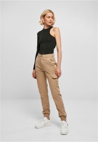 Svart top med én skulder kombinert med beige cargo-bukser. Bukser med side-lommer og elastiske mansjetter, iført hvite sneakers.