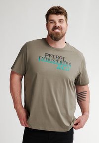 Olivově zelené bavlněné tričko s krátkým rukávem, na kterém je na hrudi vytištěno "PETROL INDUSTRIES & CO" v černé a tyrkysové barvě.