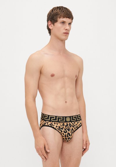 Männliches Modell mit kurzen braunen Haaren, das Leopardenmuster-Unterwäsche mit einem schwarz-goldenen Musterbund trägt, steht vor einem schlichten Hintergrund.