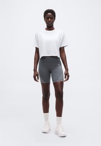 Γυναίκα που στέκεται φορώντας γκρι αθλητικές σορτς, λευκό κοντό μπλουζάκι, λευκές κάλτσες και λευκά αθλητικά παπούτσια μπροστά σε απλό ανοιχτό φόντο.