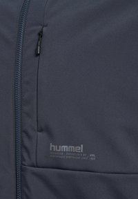 Marinblå zip-up jacka i slät tyg med en logotyp och text på vänster sida, som visar en minimalistisk design med praktiska inslag.