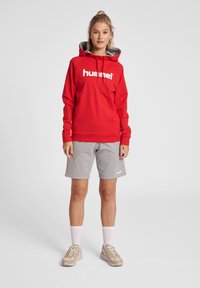 Sweat à capuche rouge avec des accents gris et un logo blanc, associé à un short gris. Le modèle porte des baskets claires et des chaussettes blanches.