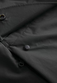 Mørkegrå stof med en subtil tekstur, der har en sort trykknap og lynlåsdetalje. Materialet ser let og glat ud.