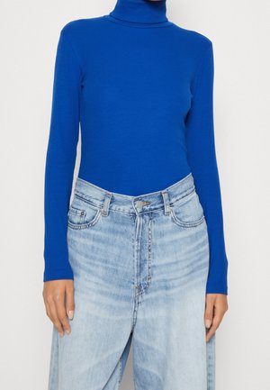 Personne portant un col roulé à manches longues bleu vif rentré dans un jean taille haute délavé clair, les mains détendues le long du corps.