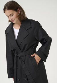 Cappotto trench nero con ampi rever, bottoni doppi e cintura in vita legata. Presenta una texture liscia e cinturini regolabili sui polsini.