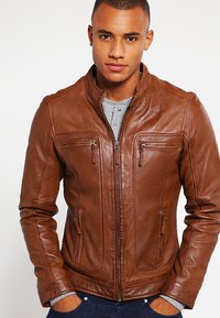 Chaqueta de cuero marrón con cuello alto, bolsillos con cremallera y un diseño ajustado; cuenta con acentos cosidos y una textura suave.