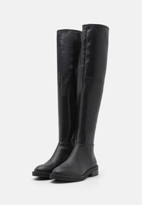 Botas de cuero negro hasta la rodilla con una textura suave, puntera redondeada y cremallera lateral. Suelas planas y un detalle de costura mínima en la parte superior.
