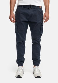 Man wearing navy blue cargo jogger pants with elastic cuffs and white sneakers, standing with hands relaxed by sides.