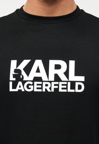 Μαύρο t-shirt με έντονο λευκό κείμενο "KARL LAGERFELD", που παρουσιάζει τη σιλουέτα ενός ατόμου με γυαλιά ηλίου και αλογοουρά μέσα στο γράμμα K.