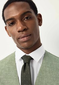 Green textured vest over a white shirt, paired with a dark green tie featuring small white polka dots. Close-up of facial features.