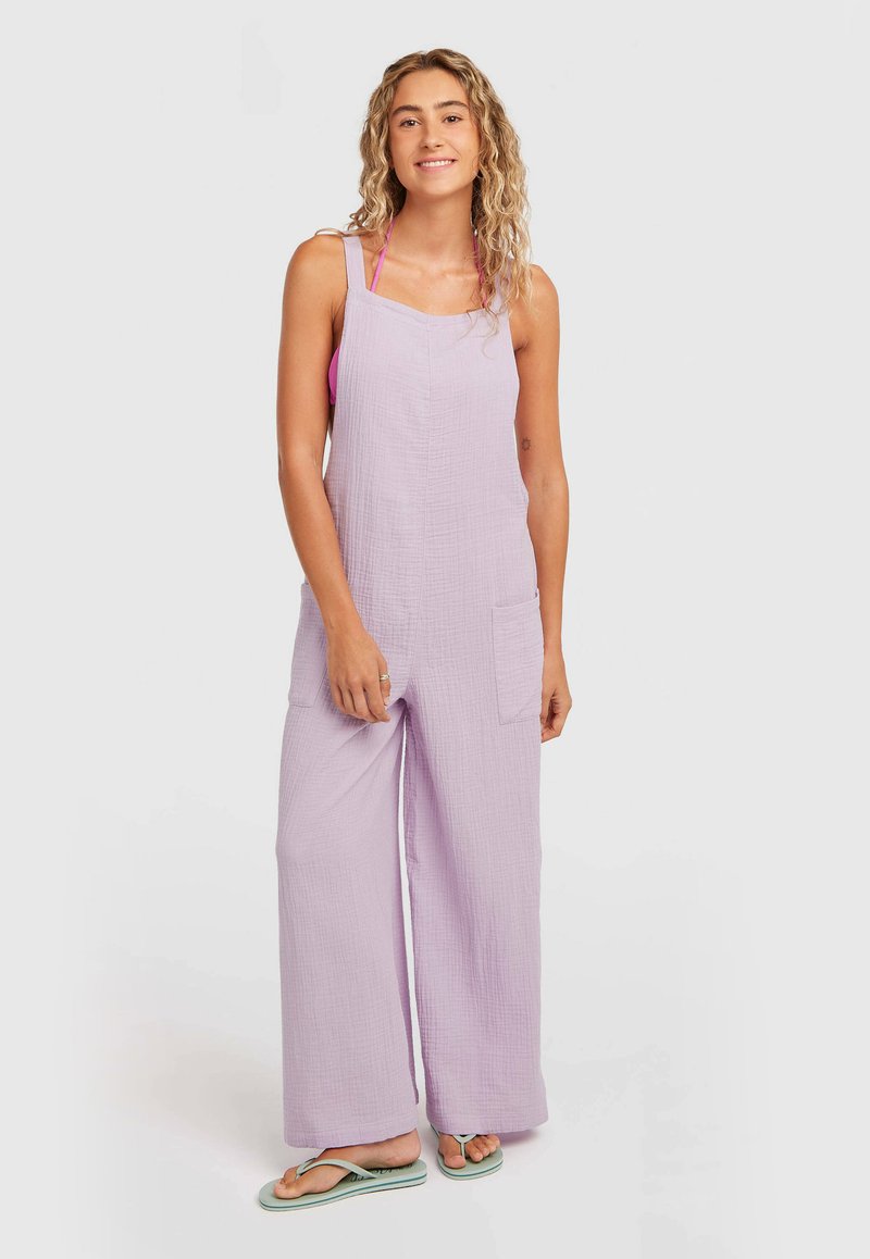 Salopette lavande en mélange de lin à jambes larges, avec deux poches avant et bretelles réglables. Portée avec un haut rose et des tongs.