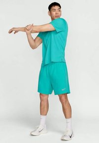 Hombre en un traje deportivo turquesa estirando su brazo derecho cruzado sobre el pecho, de pie sobre un fondo blanco y vistiendo zapatillas blancas.