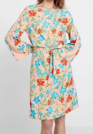 Femme portant une robe rose pâle à fleurs bleues et rouges, avec de longues manches bouffantes et une ceinture assortie en tissu nouée à la taille.