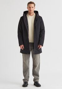 Schwarze wasserdichte Parka mit Kapuze, Frontreißverschluss und Seitentaschen. Getragen über einem cremefarbenen Pullover und hellgrauen Jeans, kombiniert mit schwarzen Stiefeln.